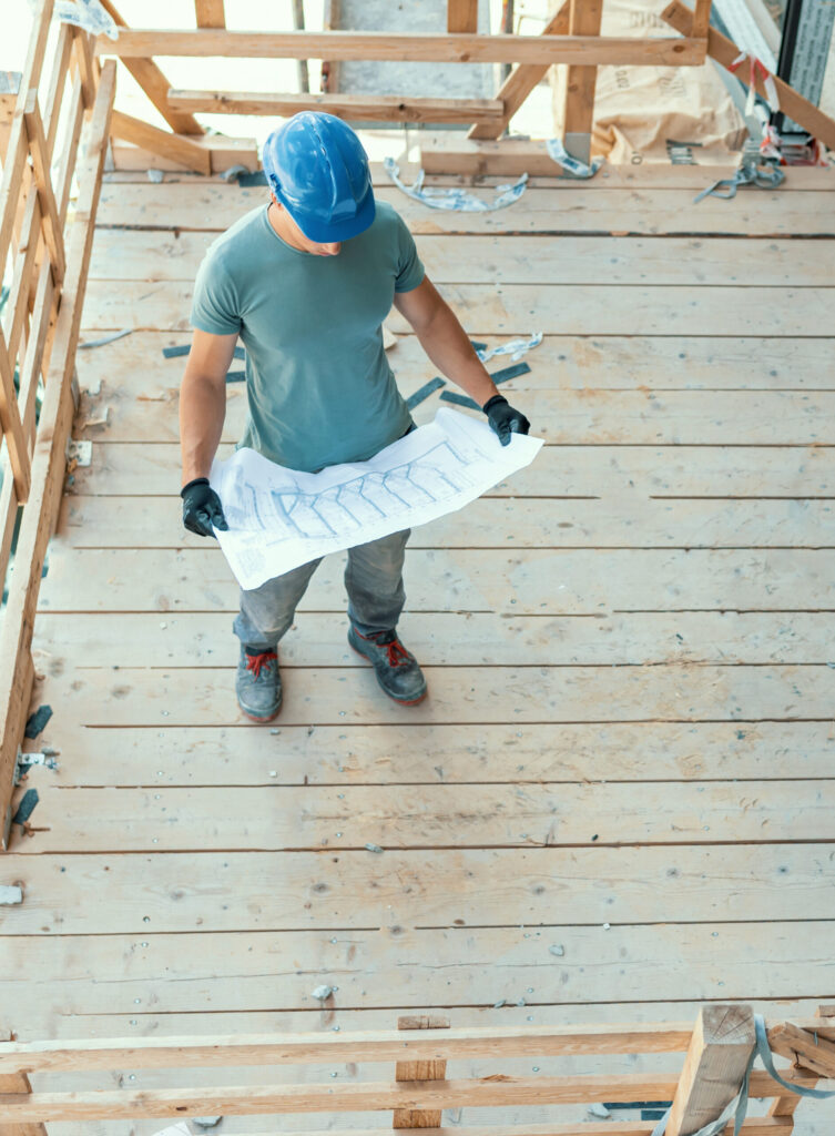 Ein junger kaukasischer Bauarbeiter steht auf dem Balkon eines im Bau befindlichen Gebäudes und hält die Baupläne in den Händen.